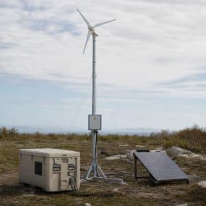 Une petite éolienne modulaire montée sur un mât sur un site isolé, avec des caisses de stockage d'énergie à proximité.