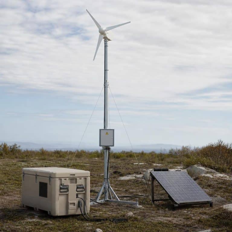 Une petite éolienne modulaire montée sur un mât sur un site isolé, avec des caisses de stockage d'énergie à proximité.