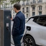 Main d'automobiliste devant une borne de recharge publique avec smartphone et QR code visible